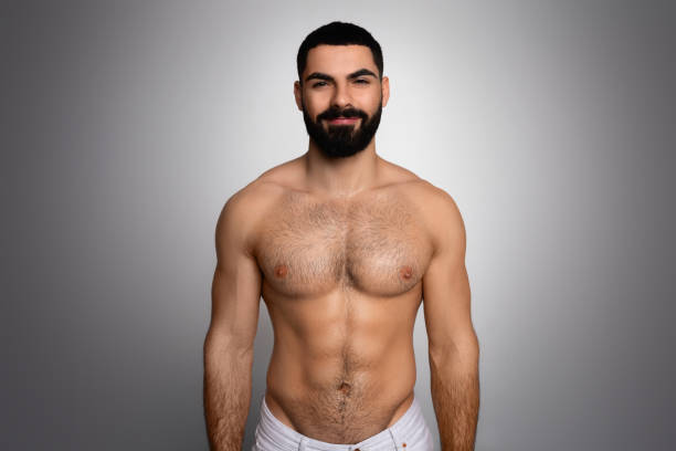 Studio shot of handsome athletic man posing after shower Studio shot of handsome young middle eastern bearded athletic man posing on grey background after shower, wrapped in bath towel, smiling at camera, showing his muscular body man muscular chest stock pictures, royalty-free photos & images