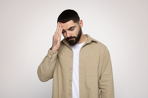 Man with a beard feeling stressed or having a headache, holding his forehead with a pained expression, dressed in a beige shirt and white tee, on a soft background, studio