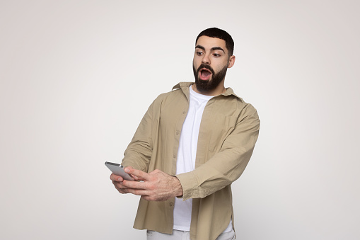Shocked millennial arab bearded man with wide-open mouth, wearing a beige shirt and white tee, excitedly looks at his smartphone, possibly reacting to surprising or amazing news