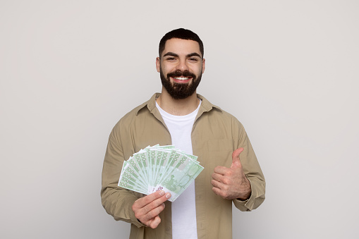 Smiling millennial arab man in a beige shirt holds a fan of 100-euro bills in one hand and gives a thumbs up with the other, suggesting financial success or a profitable deal