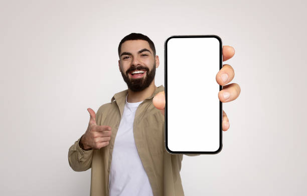 istock Enthusiastic young man with a beard, in a khaki shirt, cheerfully holds a smartphone with a blank screen 2140480868