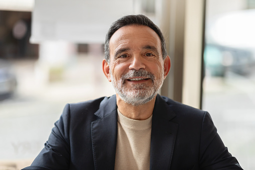 Portrait of a genial boss senior caucasian man with a neat beard, wearing a jacket and sweater, smiling warmly in a naturally lit room with a blurred background, close up