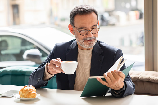 Intelligent mature man with glasses enjoying a cup of coffee while reading a book, with a delicious croissant on the side, in a comfortable cafe setting with a street view
