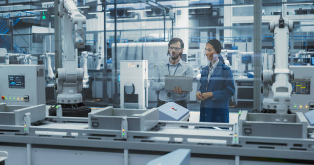 istock Two Diverse Engineers Standing in a Factory, Using Laptop at an Electronics Factory. Female and Male Specialists Chatting, Monitoring Assembly Robots on a Modern Production Conveyor Belt 2140505594