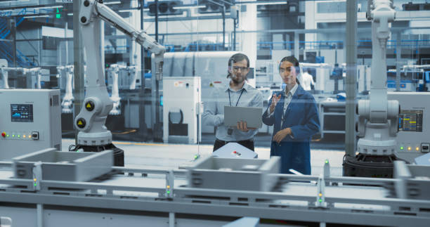 istock Two Diverse Engineers Standing in a Factory, Using Laptop Computer at an Electronics Factory. Female and Male Specialists Chatting, Monitoring Assembly Robots on a Modern Production Conveyor Belt 2140506112