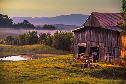 A countryside barn at dusk, the mountains in the background, a calm cow pond and rolling hills.