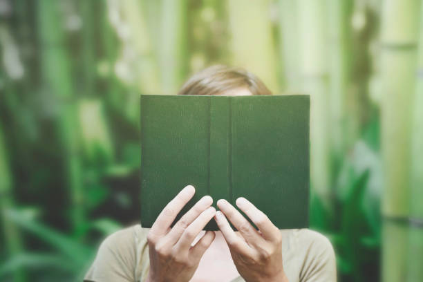surreal moment of a person with a green book in front of his face blending in with the surrounding nature and disappearing - imagination fantasy invisible women imagens e fotografias de stock