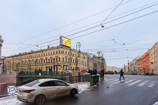 St. Petersburg, Russia, February 10, 2024. The intersection of the river embankment with the central avenue, wide roads, traffic, pedestrians and a bridge, the facade of an ancient house in the classicist style with a modern advertising screen, city landscape, city life, cars, winter.