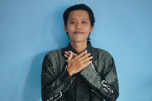 Portrait of happy asian male wearing batik shirt standing while holding his chest. He feels relieved