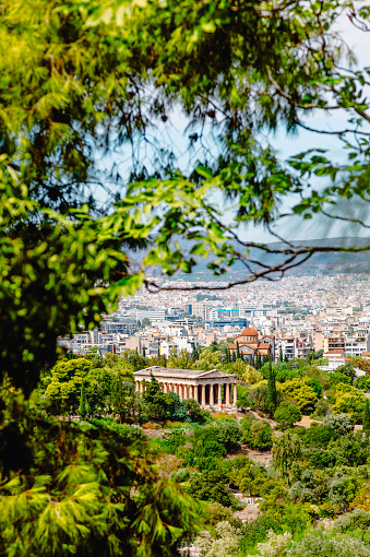 Aerial city view of the vibrant springtime landscape of Athens, Greece, as captured from the Areopagus Hill. Below, the majestic Temple of Hephaestus graces the ancient Agora, its marble columns standing proud amidst lush greenery. Against the backdrop of the bustling city, white buildings and ancient landmarks emerge, blending harmoniously with the verdant trees of spring. From the elevated viewpoint of the Acropolis, the cityscape is framed by a canopy of fresh green leaves, offering a picturesque tableau of Athens' timeless beauty and historical significance