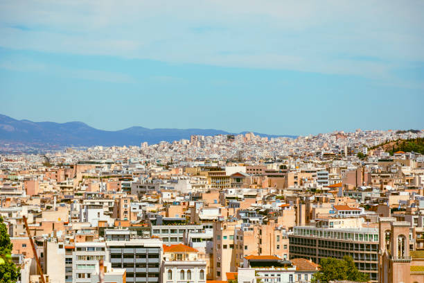 Gaze upon the modern Athenian cityscape from the elevated vantage point of the Acropolis, where the ancient and contemporary seamlessly intertwine. Splitting the central district, a wide avenue cuts through the urban landscape, symbolizing the city's evolution over millennia. Against the backdrop of a brilliant sunny sky, towering skyscrapers and historic landmarks punctuate the horizon, reflecting the dynamic spirit of Athens, Attica, Greece. From the timeless ruins of the Acropolis to the bustling streets below, this panoramic view captures the essence of Athens' enduring allure in the warm embrace of sunlight