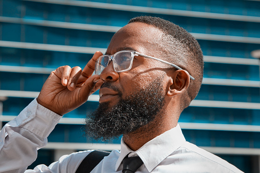 A professional portrait of a black businessman with a beard, adjusting his glasses against the backdrop of a modern cityscape. This image exudes confidence, ambition, and sophistication in a corporate environment.