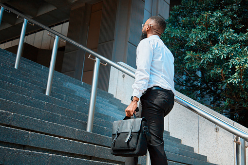 A black businessman in a white shirt and carrying a briefcase ascends outdoor stairs, exuding confidence amidst a bustling city environment.