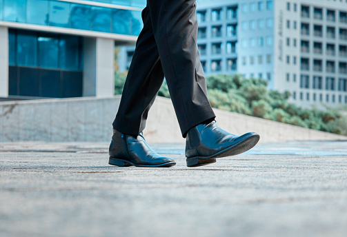 A dynamic low-angle view capturing the stride of a black businessman dressed in a sharp suit. He confidently walks through a cityscape, symbolizing ambition, success, and corporate lifestyle.