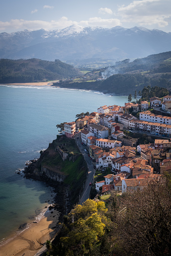 Vertical shot reveals Lastres coastal charm, with vibrant sea and snowy mountains.