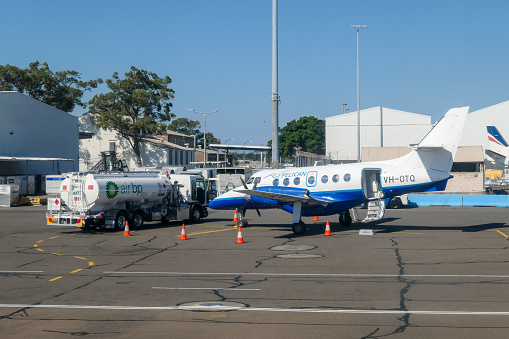 A refueling truck has pulled up alongside a FlyPelican BAe Jetstream 32 plane, registration VH-OTQ, parked on the tarmac of Sydney Kingsford-Smith Airport.  In the right background is the vertical stabiliser of a Rex Airlines Saab 340B plane. This image was taken from a Virgin Australia B737-81D plane, registration VH-YFC, taxiing to the third runway for departure as flight VA850 to Melbourne on a sunny afternoon on 1 April 2024.