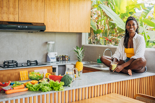 Black woman enjoys a peaceful moment in a modern, sunlit kitchen. She is seated on the countertop, holding a glass of smoothie.