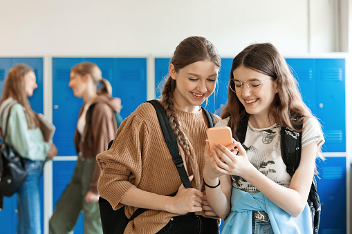 Two happy female high school students standing in school corridor against blue lockers and using mobile phone.