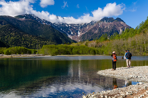 Hikers next to a pond and the Azusa River with snow-capped mountains behind. Kamikochi, Nagano Prefecture