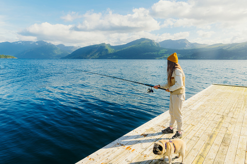 Side view of of female-fisherman with long hair fishing with her pug at the wooden pier with scenic view of the sea and green mountain peaks during sunny summer day in Norway Fjords