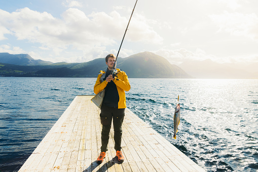 Front view of a smiling male fisherman happy to catch fish by fishing rod at the wooden pier with scenic view pf the sea and green mountain peaks in Norway