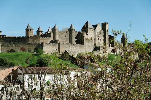 Horizontal landscape photo of the cobblestone paved walkway leading to the former drawbridge entrance in the surrounding wall of the castle at Saumur. Trees, Castle towers and rooftops can be seen in the distance under a blue sky. Saumur, Maine-et-Loire, France. 1st April, 2019