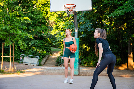 Two women playing basketball in a park. One of them is holding a basketball. The other woman is wearing a green tank top