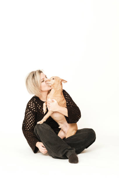 Blonde woman kissing her brown hound seated in a white background