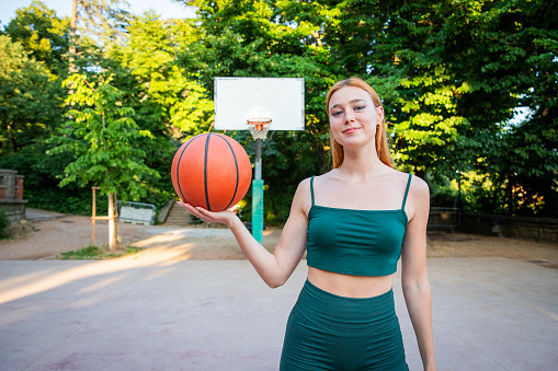 A woman is holding a basketball in a park. The park is surrounded by trees and has a basketball court. The woman is smiling and she is enjoying herself