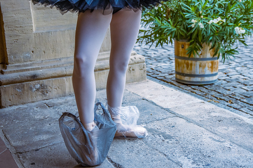 Ballerina standing on the stone floor wearing pointe shoes with black and white plastic bag protecting shoes. Ballet dancer with white tights on. Backstage. Street performance. Concept. Funny photo