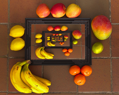 Droste effect (mise en abyme or recursive) shot of fruit (bananas, lemons, lime, clemantines, appples, mango) with a picture frame on a tiled stone floor