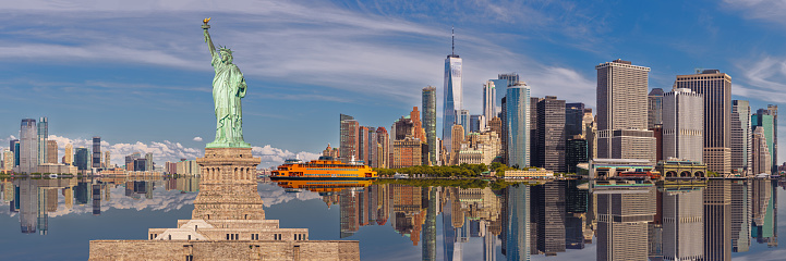 Statue of Liberty and New York City Skyline with Jersey City, NJ, Manhattan Financial District, Battery Park, World Trade Center, Staten Island Ferry, Blue Sky with Clouds all Reflected in Water of New York Harbor. High Resolution Stitched Panoramic image with 3:1 image aspect ratio. Canon EOS 6D Full Frame Sensor Camera and Canon EF 85mm f/1.8 USM Prime Lens.