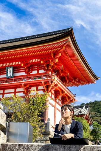 Kyoto, Japan - October 10th, 2023: A Close-up view of a business woman overlooking the views of Kyoto from the grounds in front of a famous red Pagoda under a blue autumn sky in Kyoto Japan.