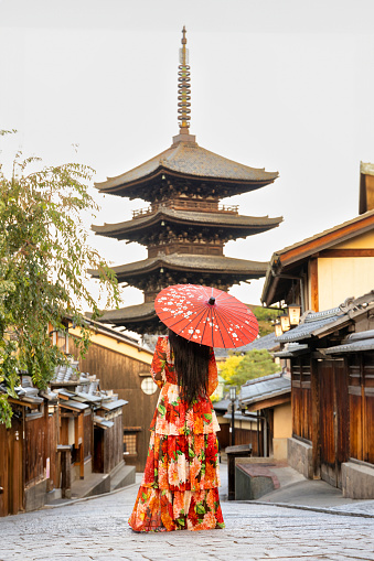 Kyoto, Japan - October 11, 2023: A tourist standing in the famous Sannen Zaka street in the Gion district in Kyoto, Japan, wearing a red dress and holding an umbrella and Yasaka Pagoda in the background.