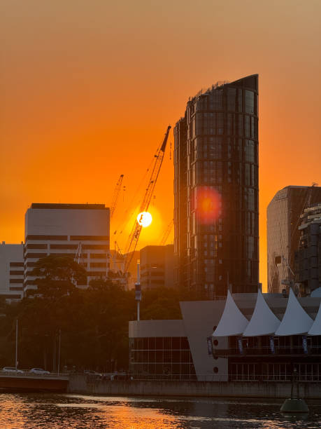 A city skyline with a large building in the foreground and a crane in the background. The sun is setting, casting a warm glow over the scene