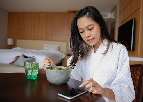 Close-up shot of Young Indonesian Woman Buying Cryptocurrency For Investments. She's using her smartphone to buy stable coin, while she's eating healthy green salad at hotel room. concept : generation z checking her crypto investment