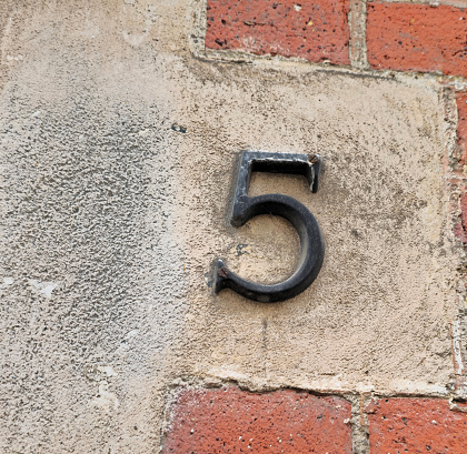 Number 5 made of metal, held on by old rusty bolts. Against the background of a brick wall, which forms some texture