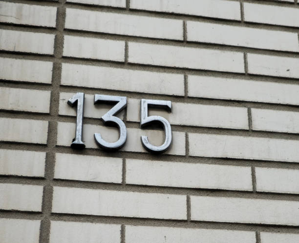 Number plate. Number (135) one hundred thirty-five against a white brick wall background.