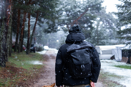 Person walking with dog through snowy campground surrounded by forest, wearing black coat, backpack