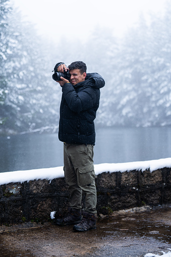 Latinx young man in black coat captures snowy forest lake with professional camera.