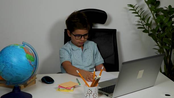 istock handsome smiling boy in glasses waves his hand while looking into laptop webcam while sitting at white table. Online learning 2141305221
