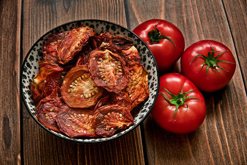 Sun dried tomatoes in bowl and three fresh ripe tomatoes on dark wooden table background. Top view.