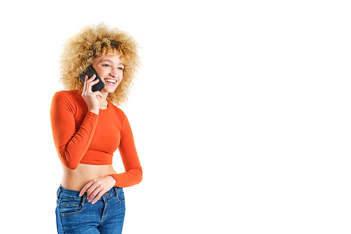 young brazilian girl with blonde afro hair smiling talking on cell phone on white background wearing orange sweater and blue jeans