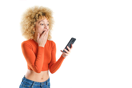 young brazilian girl with blonde afro hair with surprised expression looking at her smart phone wearing orange sweater and blue jeans on white background