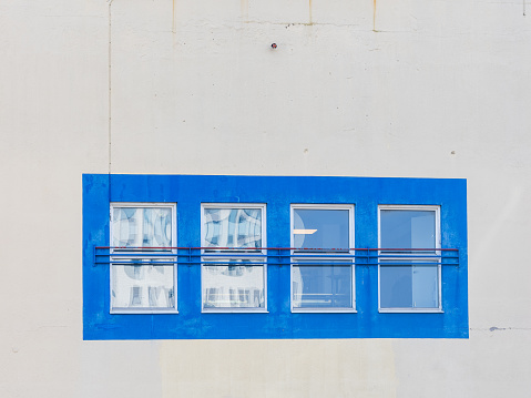 A row of four windows set within bright blue frames contrasts starkly against the minimalistic pale facade of a building. Each window reflects a different scene, with one showcasing a partially open curtain, adding a lived-in touch to the urban architecture.