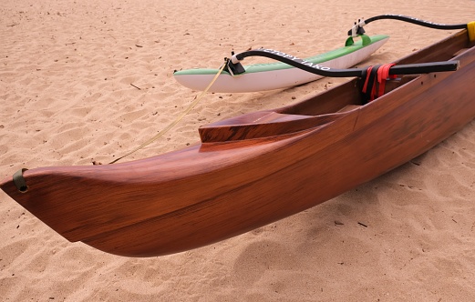 Hawaiian outrigger canoes sitting on the grass at a beach park and also sitting on the sand at the beach.  Canoes are used in a competitive sport in Hawaii and also as a relaxing way of getting exercise.  Each canoe can be different colors and made with different materials.