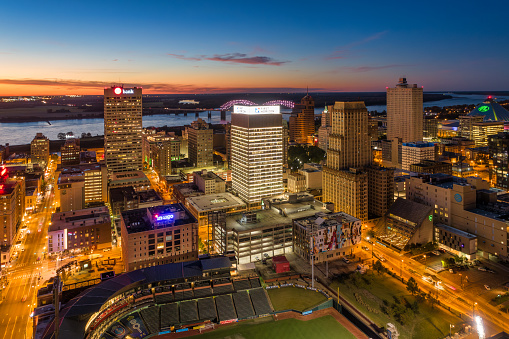 Downtown Memphis skyline aerial, with illuminated highrise buildings and streets in the foreground, and the Mississippi River, Hernando De Soto Bridge, Arkansas landscape, and a sunset painted sky in the background.