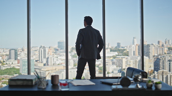 Manager silhouette contemplating city in panoramic office rear view. Calm company ceo in suit thinking looking window alone. Successful professional silhouette dreaming pondering future at workplace.