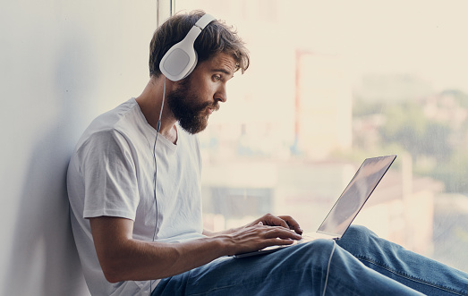 bearded man with an open laptop sits on the windowsill and headphones on his head. High quality photo