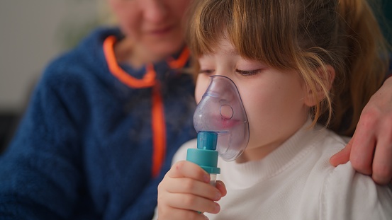 A mother helping is her daughter using a nebulizer in the living room at home.